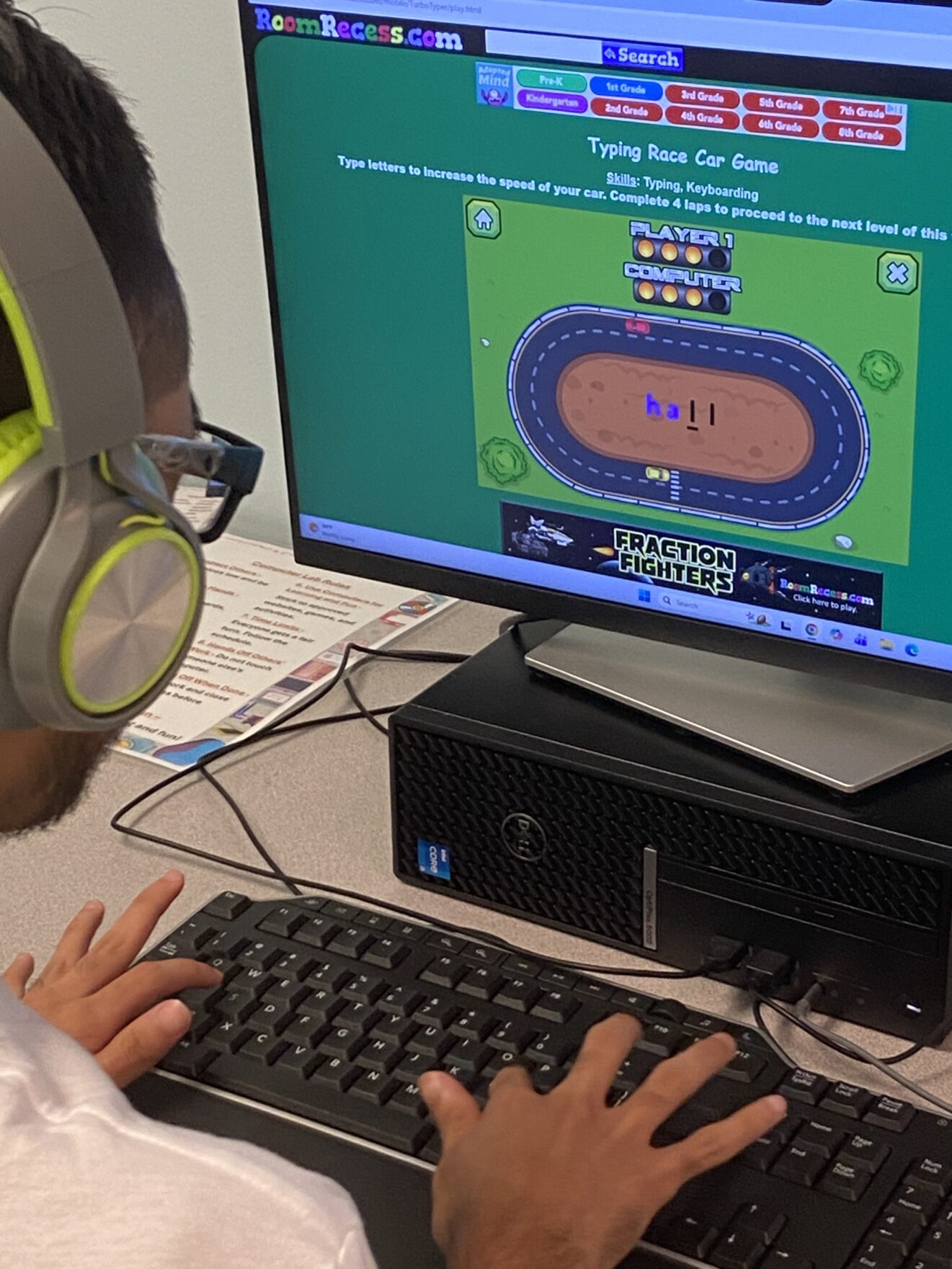 Man (with dark hair and beard) sitting at computer, wearing yellow headphones and eyeglasses. Computer monitor shows a digital racetrack ("Typing Race Car Game"). At bottom of screen, dark-colored space-adventure banner for "FRACTION FIGHTERS" game.