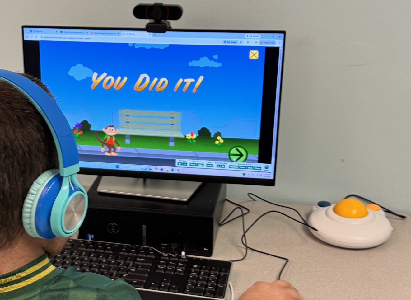 Person wearing blue headphones surveys a computer monitor displaying the results of a game. Screen image: at bottom, monkey in red shirt stands next to a gray park bench. Other park-play images dot background. In blue sky above, orange letters display the words "YOU DID IT!" 